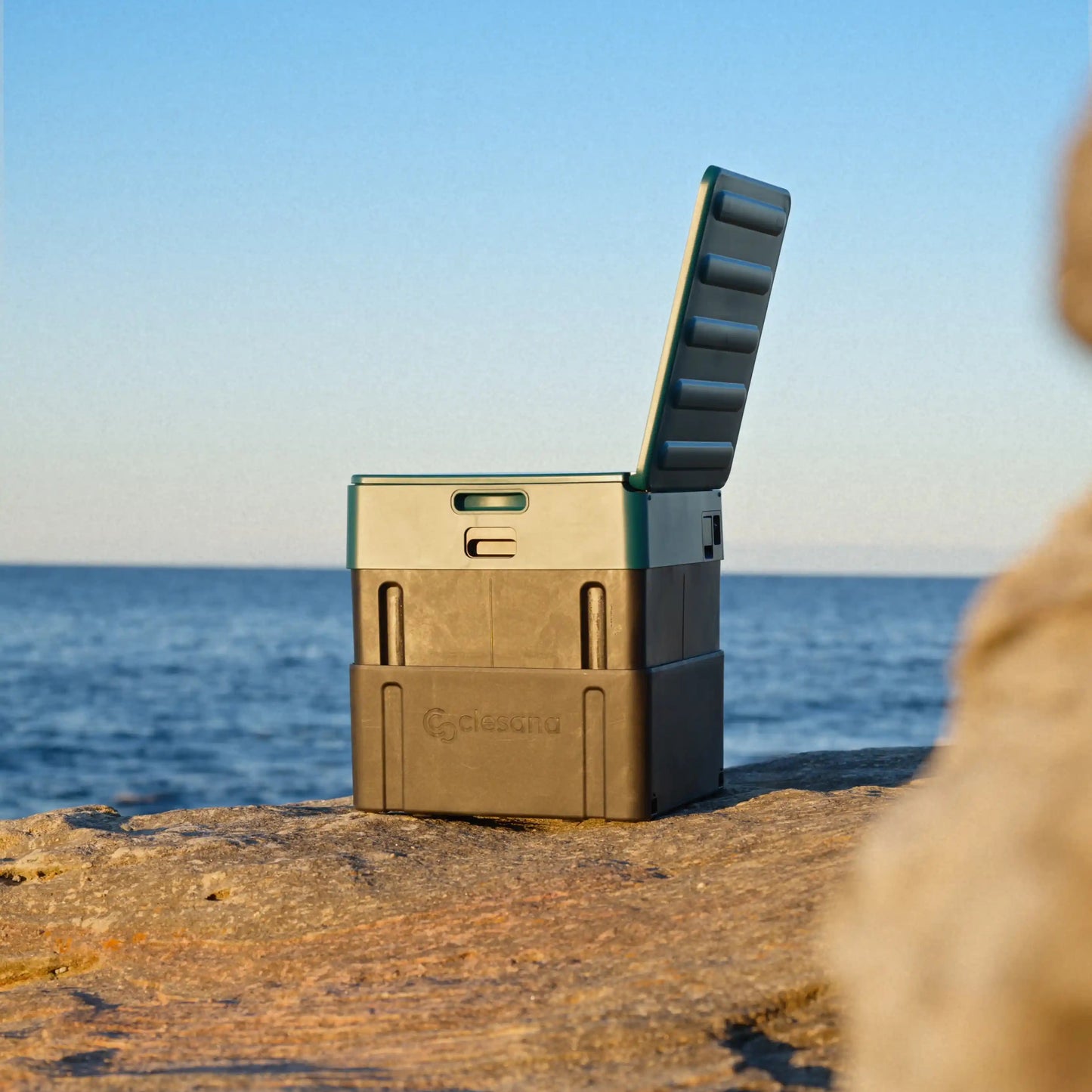 A rugged outdoor portable toilet with its lid propped open, sitting on a rocky Australian shoreline with the ocean and blue sky in the background.